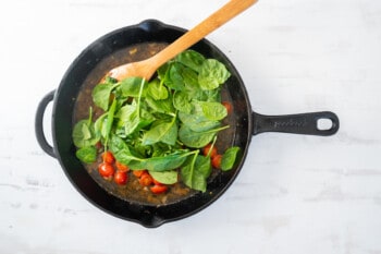 spinach added to tomato, onion, garlic, and broth in a cast iron pan with a wooden spoon.