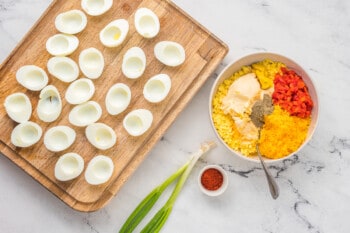 hollowed out hard boiled eggs on a wooden cutting board next to a bowl of pimento cheese deviled egg filling.