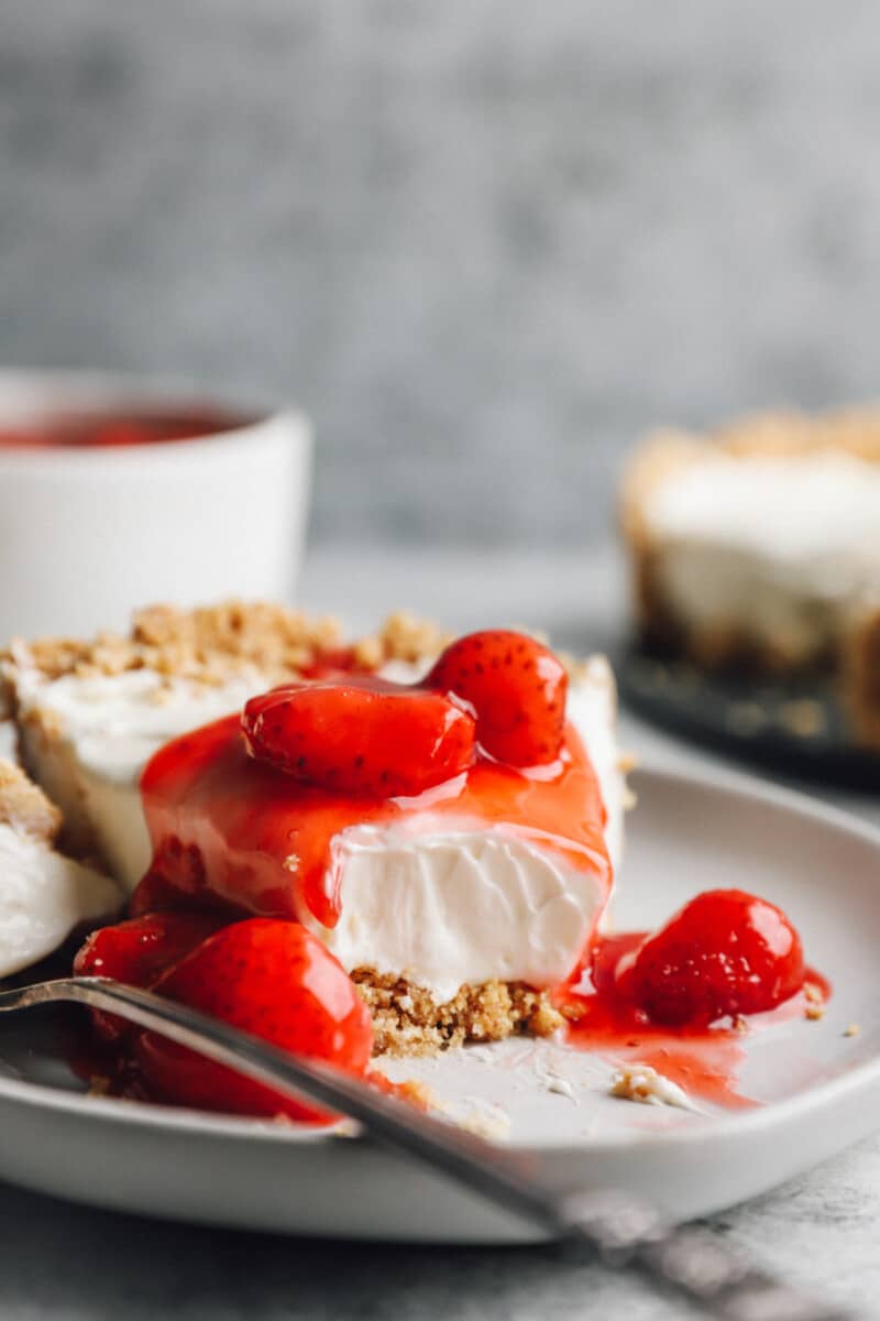 side view of a partially eaten slice of no bake cheesecake on a white plate with strawberry topping and a fork.