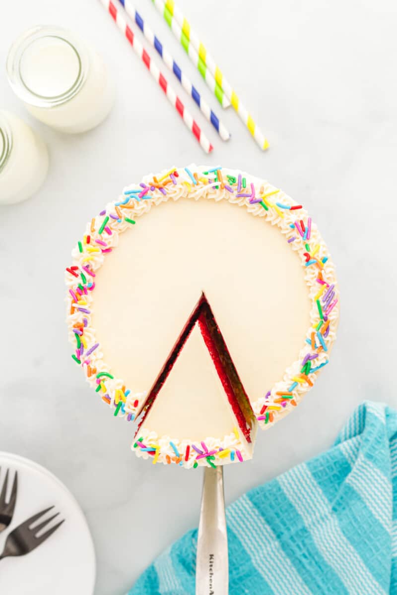 overhead view of a cake server removing a slice of rainbow cake from a rainbow cake on a white cake stand.