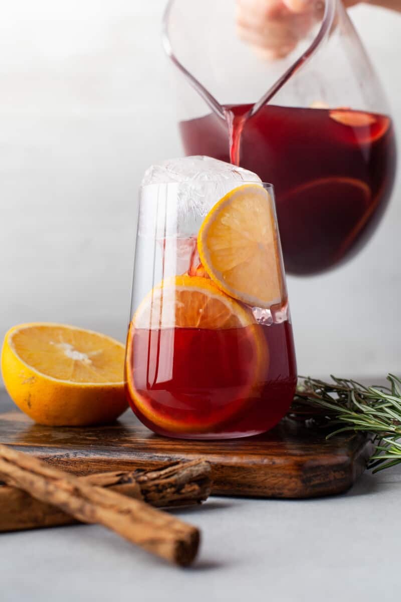 side view of red sangria in a large glass pitcher on a wooden cutting board.