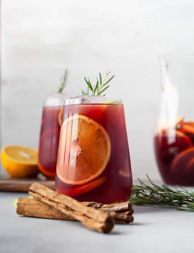 side view of cinnamon sticks crossed in front of red sangria in a stemless wine glass with ice and rosemary.
