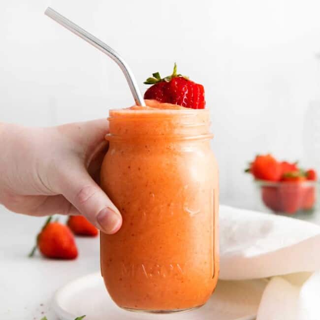 a hand grabbing a strawberry mango smoothie in a mason jar with a stainless steel straw.