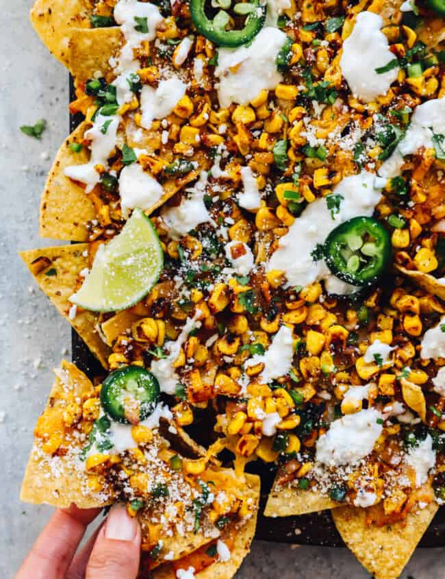 a hand grabbing from the corner of a baking sheet filled with street corn nachos.