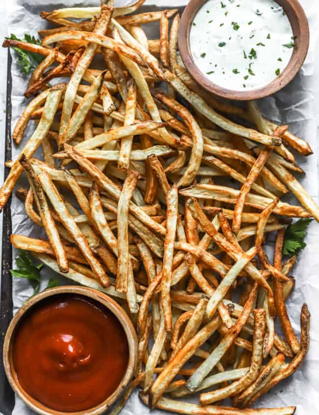 overhead view of air fryer french fries on a baking sheet with a cup of ketchup in the lower left corner and a cup of ranch in the upper right corner.
