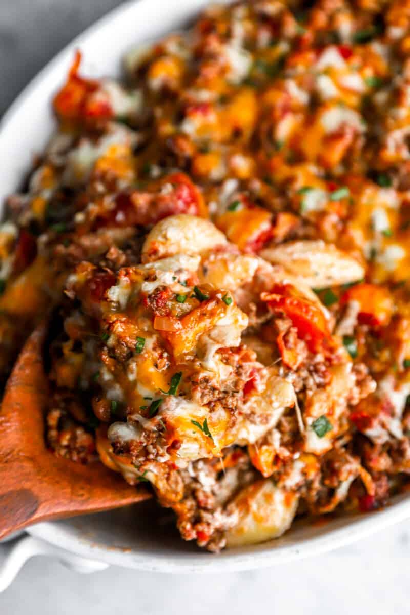 a wooden spoon lifting a scoop of hamburger casserole from a white oval casserole dish.