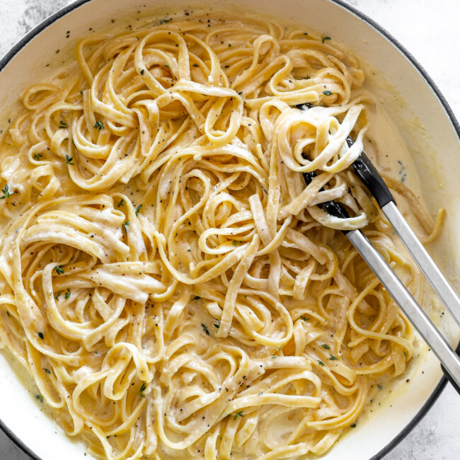 overhead view of fettuccini alfredo in a white dutch oven with tongs.