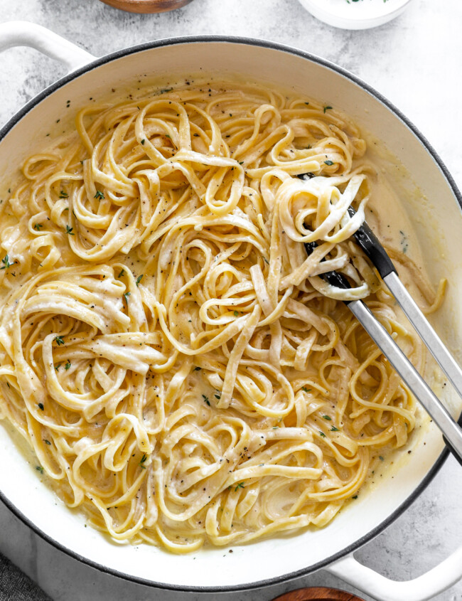 overhead view of fettuccini alfredo in a white dutch oven with tongs.