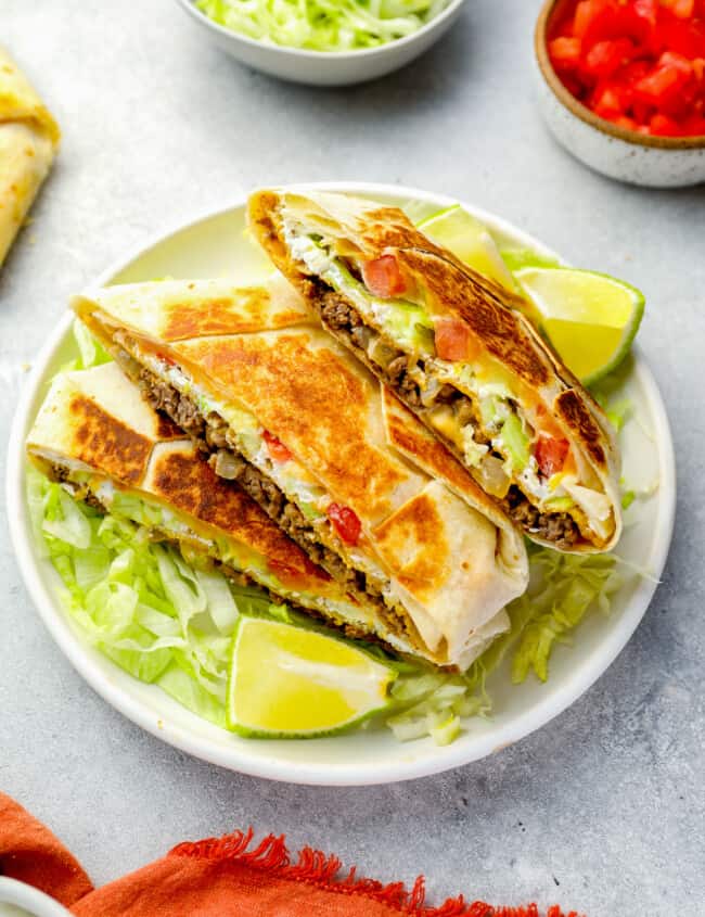 three-quarters view of a stacked halved crunchwrap supreme on a white plate with lettuce and lime wedges.