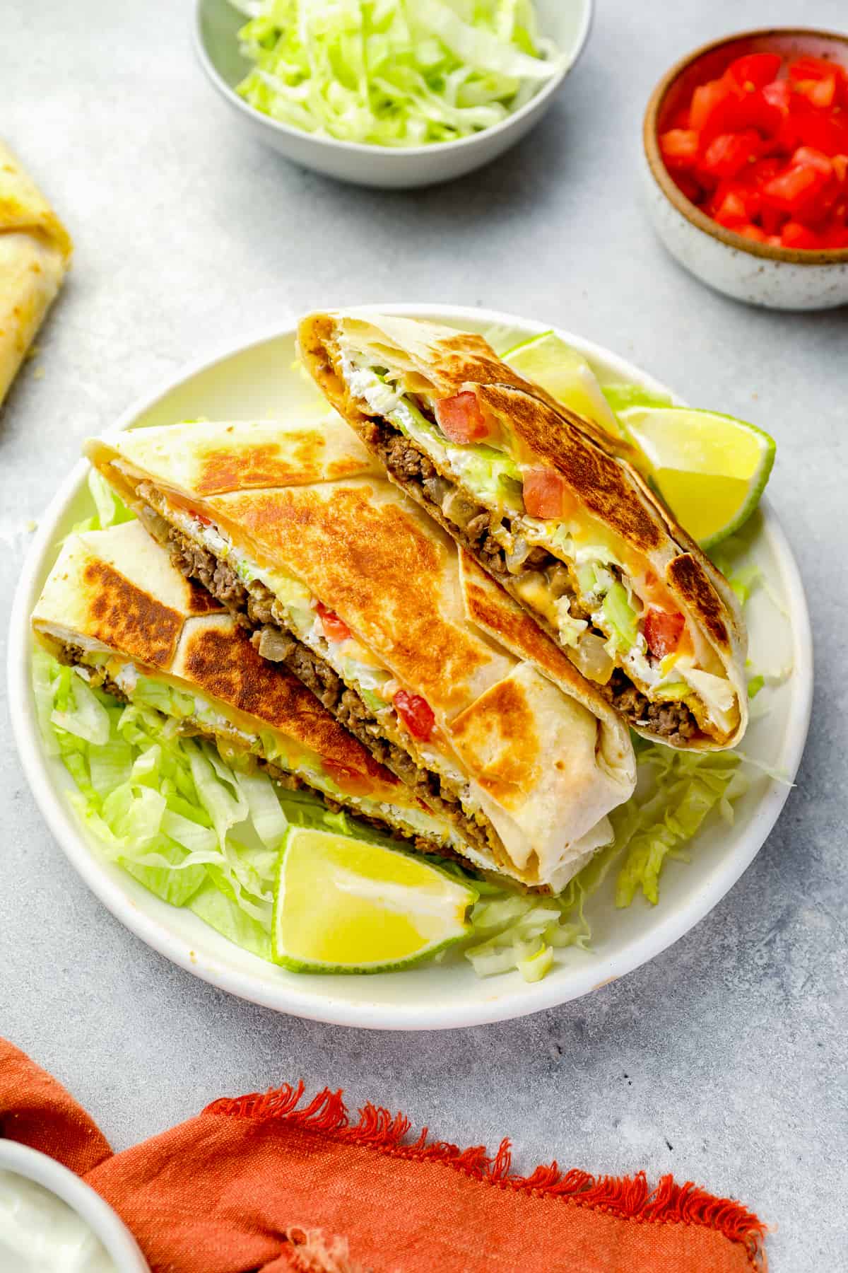 three-quarters view of a stacked halved crunchwrap supreme on a white plate with lettuce and lime wedges.