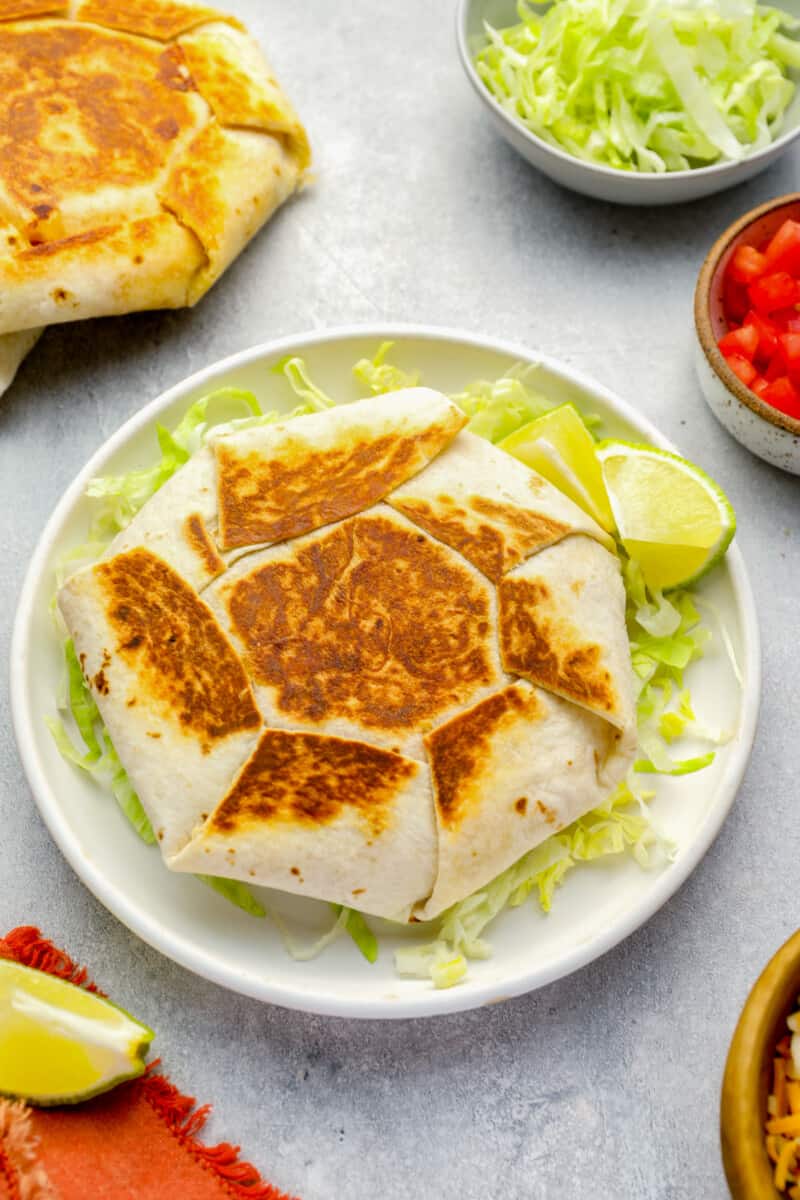 three-quarters view of a homemade crunchwrap supreme on a white plate with lettuce and lime wedges.