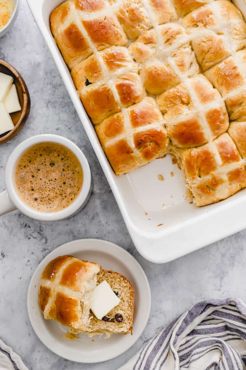overhead view of a buttered halved hot cross bun on a white plate next to a baking tray filled with buns.