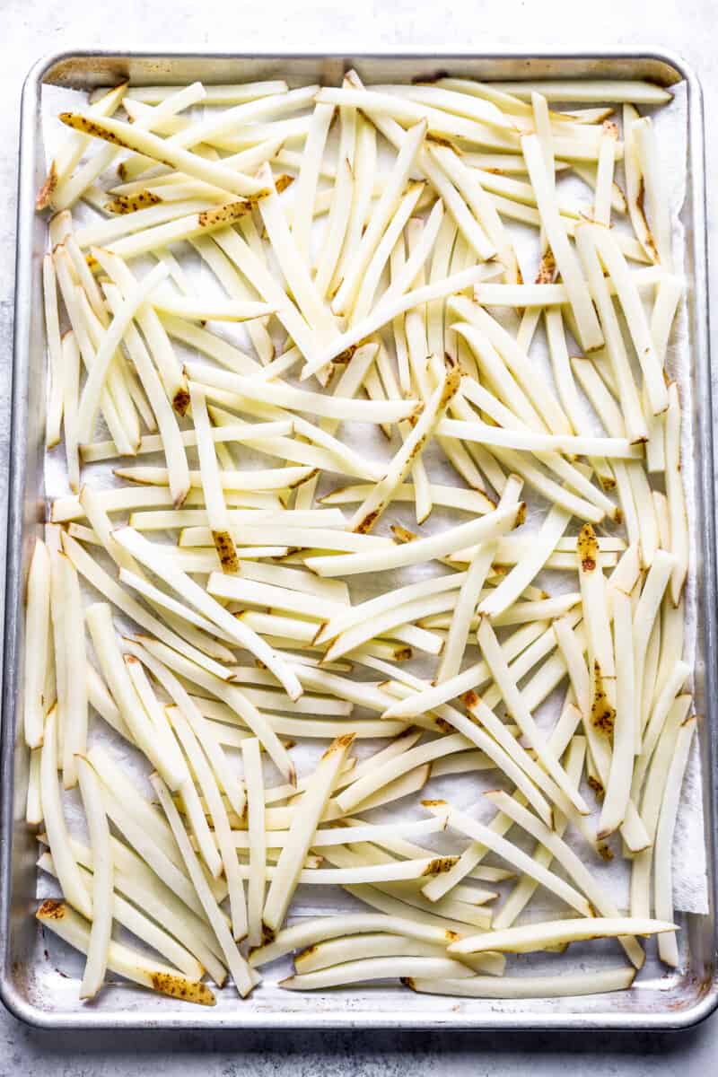 Raw French fries spread out on a baking sheet.