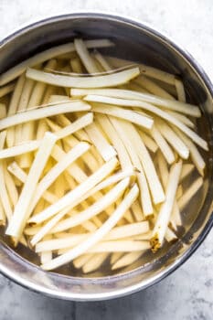 raw french fries soaking in water in a stainless steel bowl.