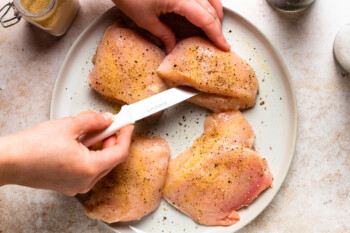 a hand using a knife to slice a pocket into 1 of 4 chicken breasts on a white plate.