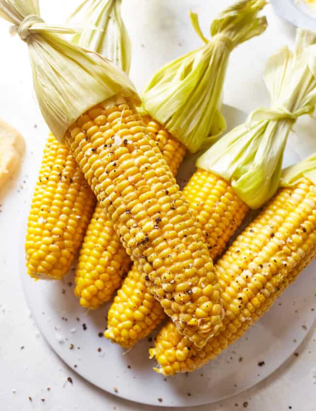 5 ears of corn on the cob with their husks tied back on a glass plate.