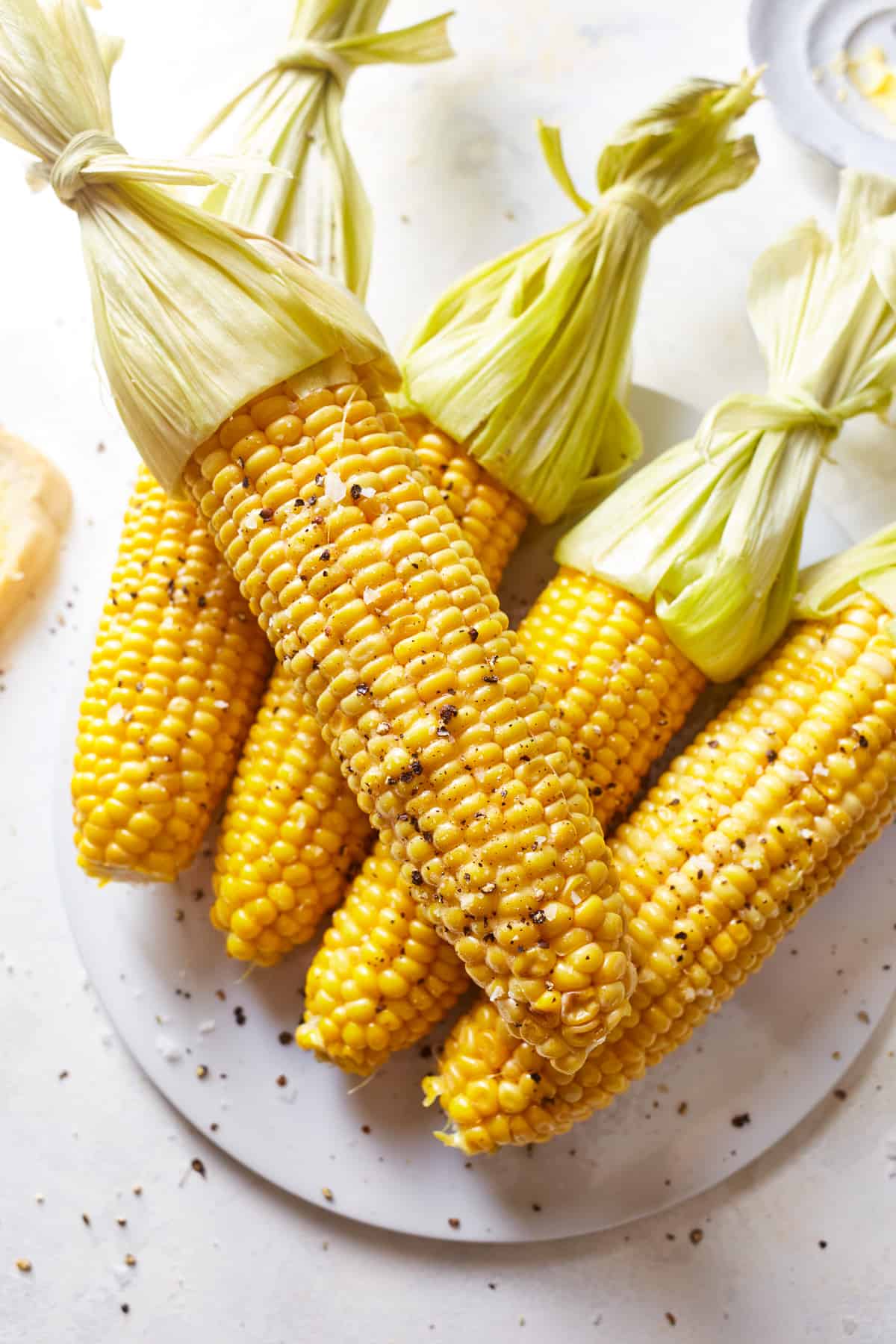 5 ears of corn on the cob with their husks tied back on a glass plate.