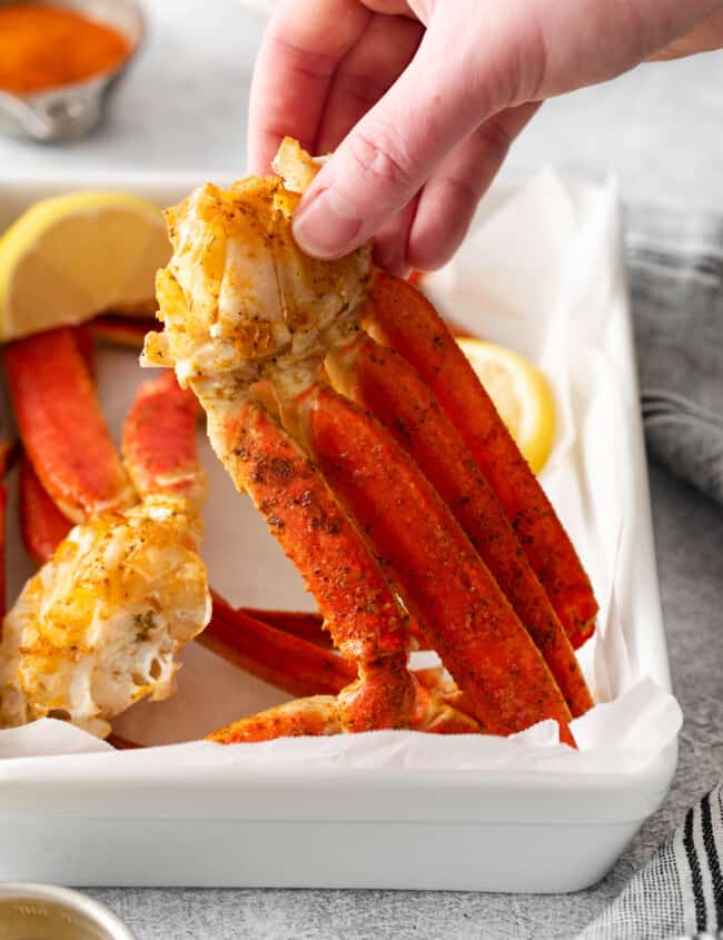 three-quarters view of a hand holding up a cluster of crab legs from a white rectangular plate.