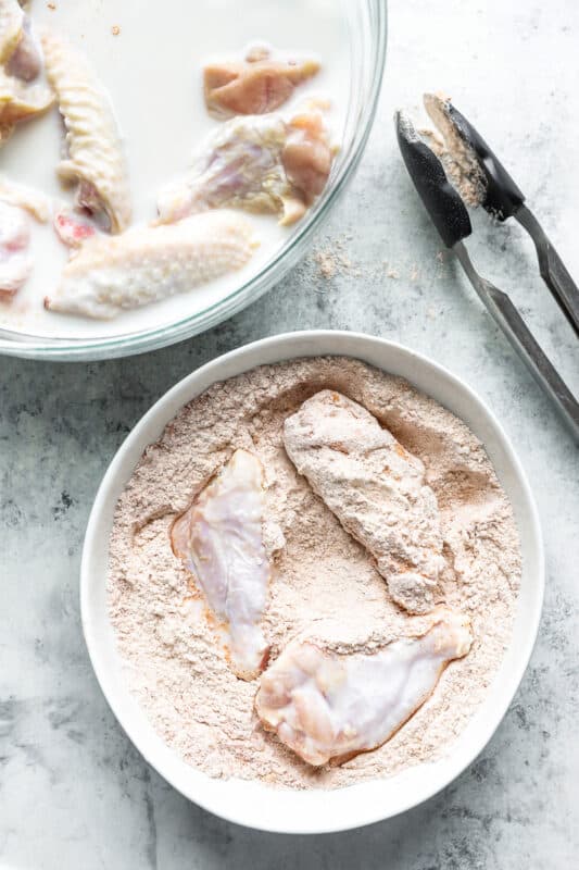 buttermilk marinated chicken wings being dredged in flour in a white bowl.