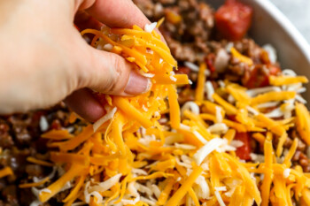 a hand sprinkling cheese over an unbaked hamburger casserole in a white casserole dish.