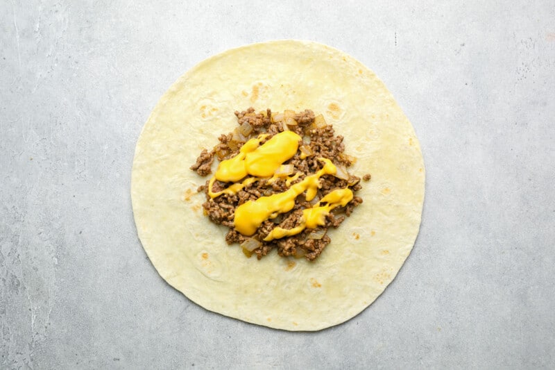 nacho cheese sauce on top of ground beef on a large flour tortilla.