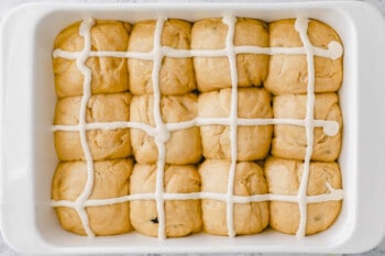 proofed hot cross bun dough balls with piped crosses on top in a rectangular baking pan.