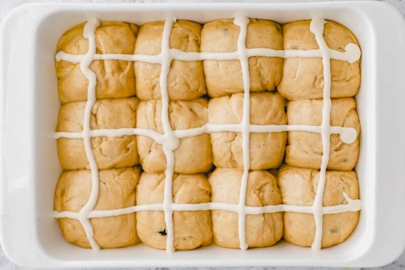 proofed hot cross bun dough balls with piped crosses on top in a rectangular baking pan.