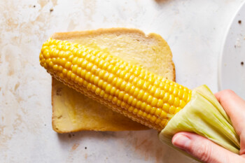 an ear of boiled corn being rubbed on a buttered slice of bread.