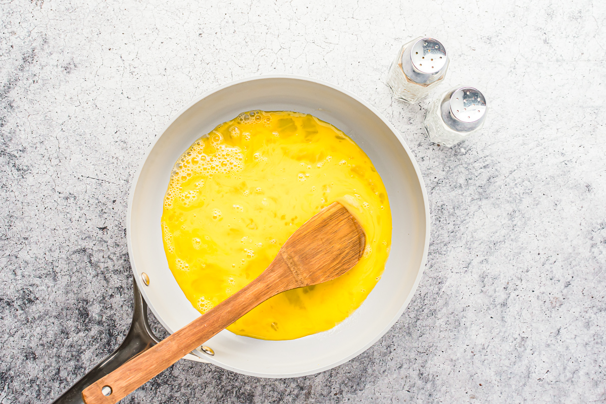 overhead view of raw eggs in a frying pan with a wooden spatula.