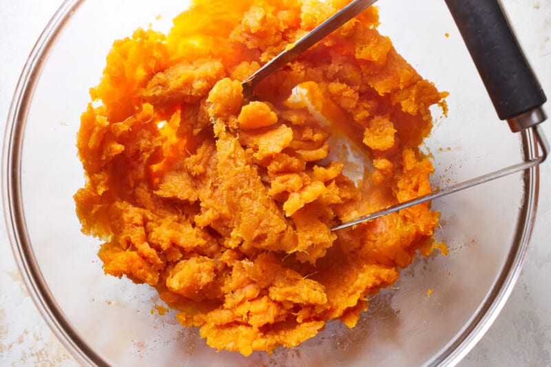 mashed sweet potatoes in a glass bowl with a potato masher.