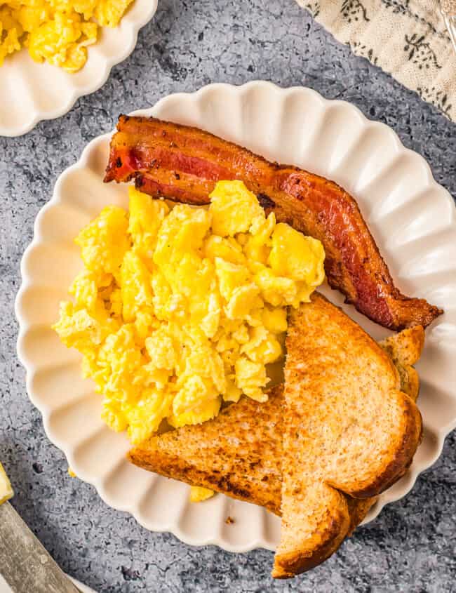 overhead view of scrambled eggs on a white plate with toast and bacon.