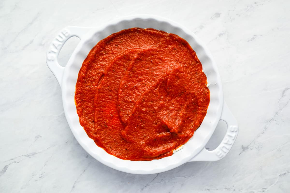 marinara sauce spread over cream cheese layer in a round white baking dish.