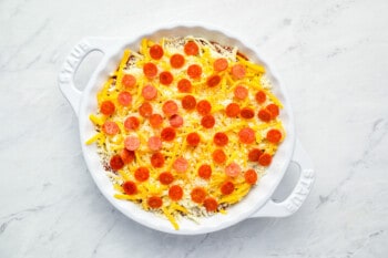 mini pepperoni sprinkled over unbaked pizza dip in a white round baking dish.