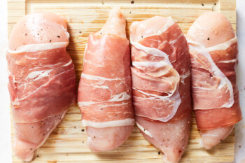 4 raw prosciutto wrapped chicken breasts on a wooden cutting board.