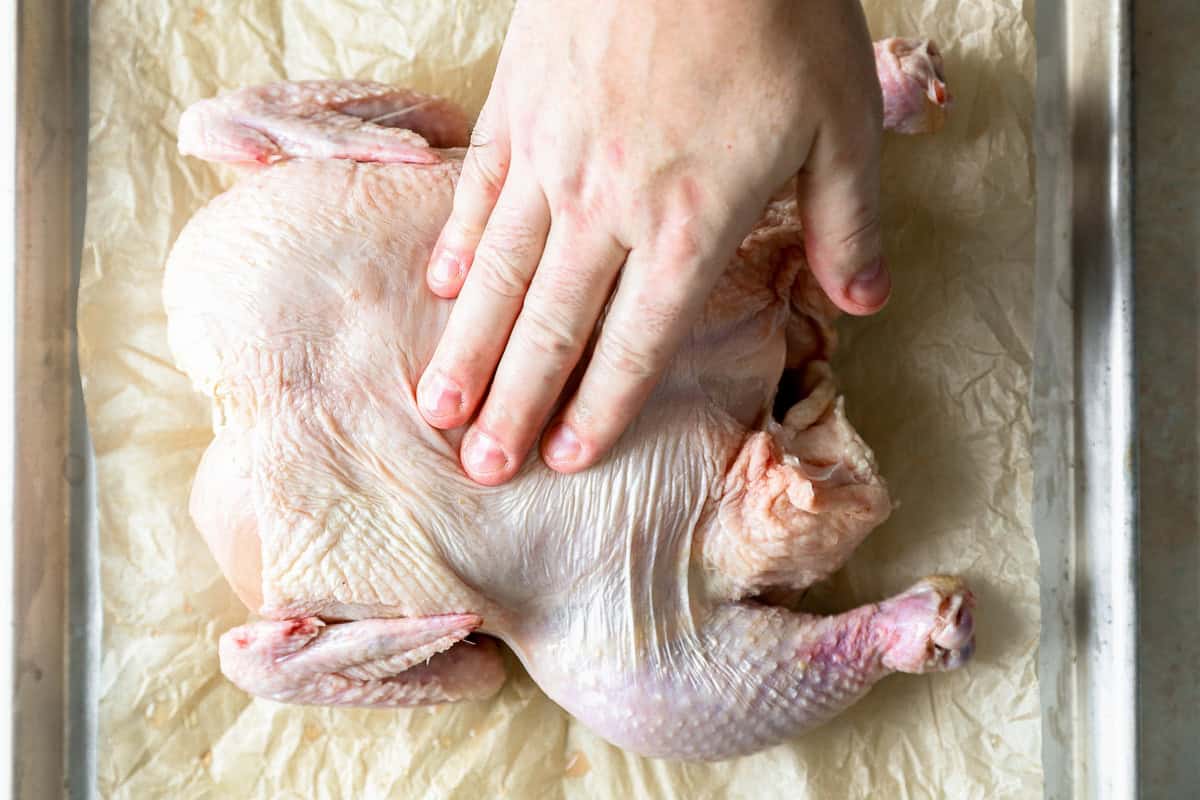 A hand pressing in the center of a whole chicken to flatten it on a parchment-lined baking sheet.