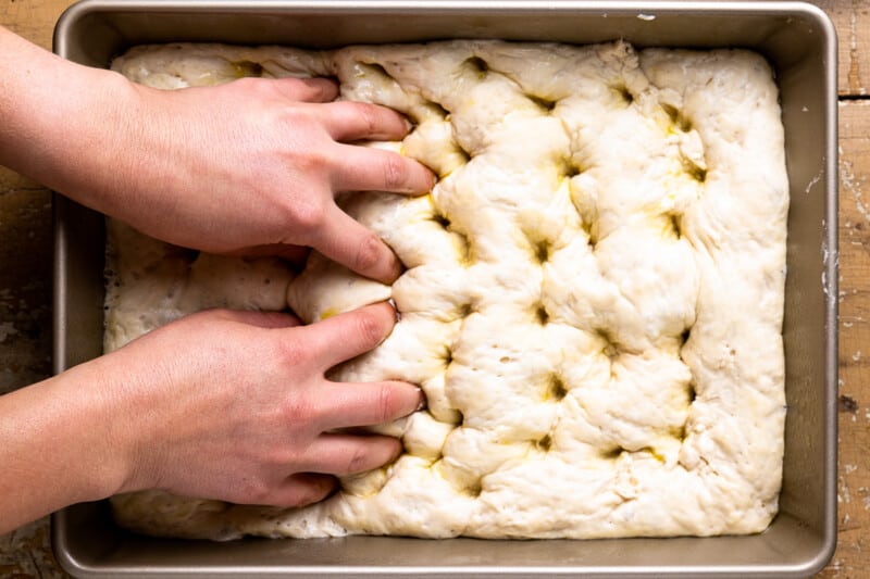 two hands using fingers to dimple no knead focaccia dough in a rectangular baking pan.