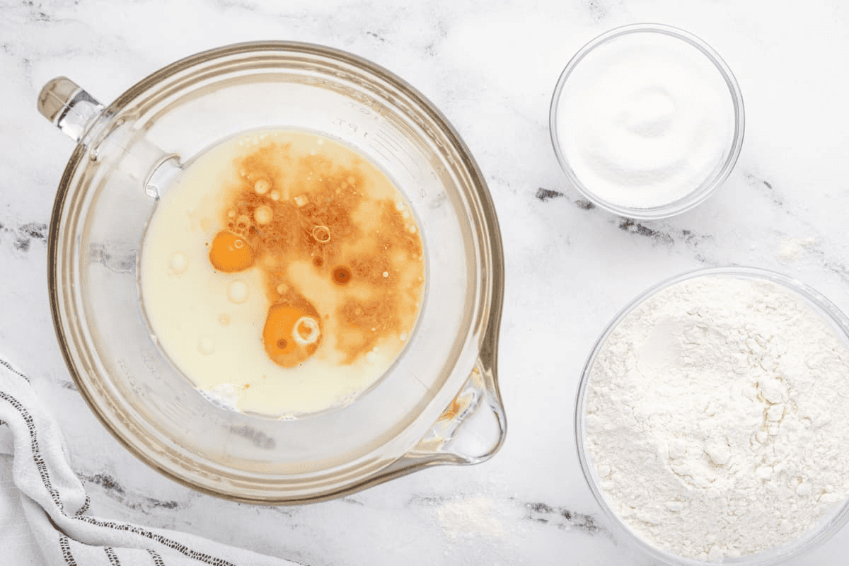 Wet ingredients in a large measuring cup with a bowl of flour and a bowl of sugar.