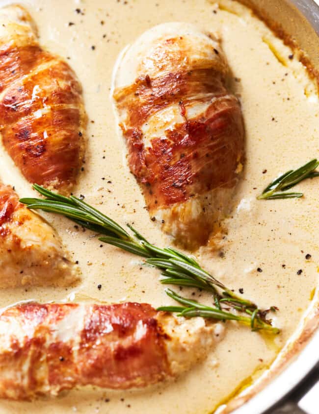 partial view of prosciutto wrapped chicken in a frying pan with rosemary.