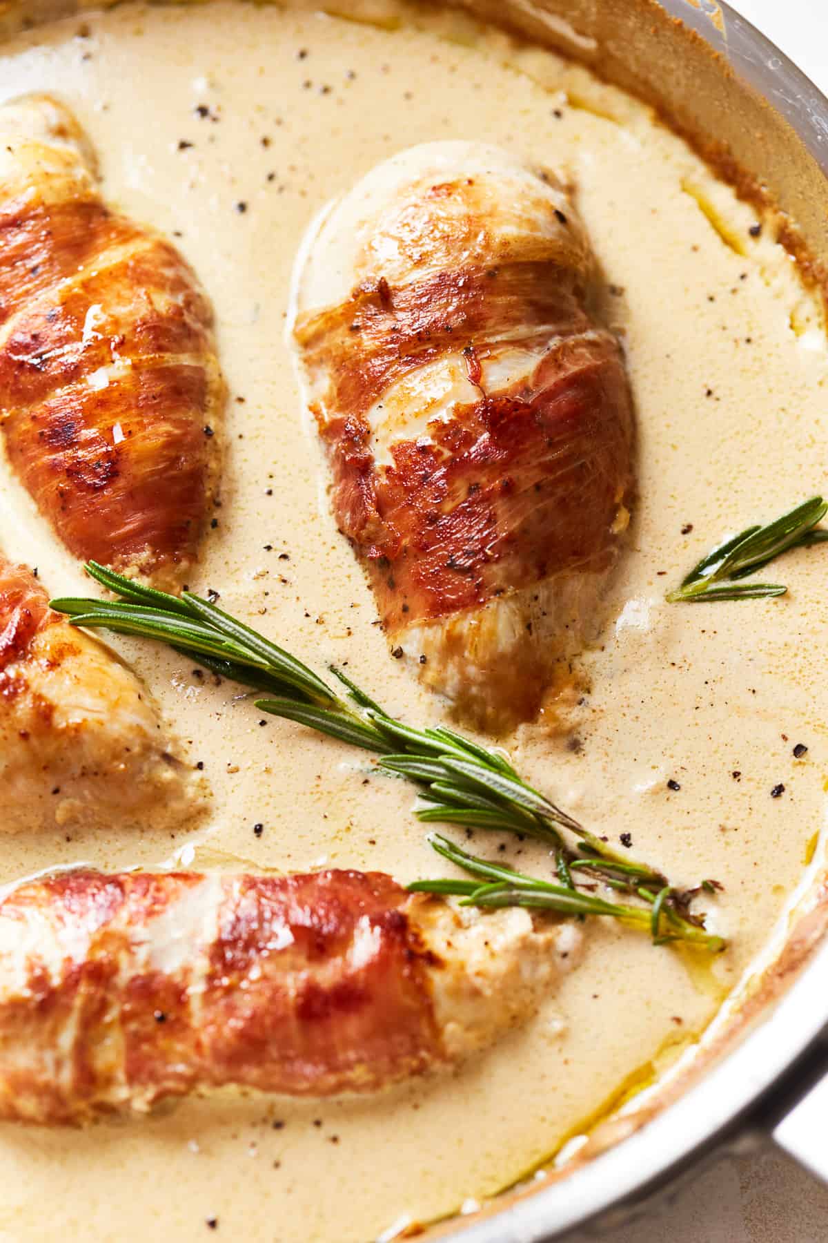 partial view of prosciutto wrapped chicken in a frying pan with rosemary.