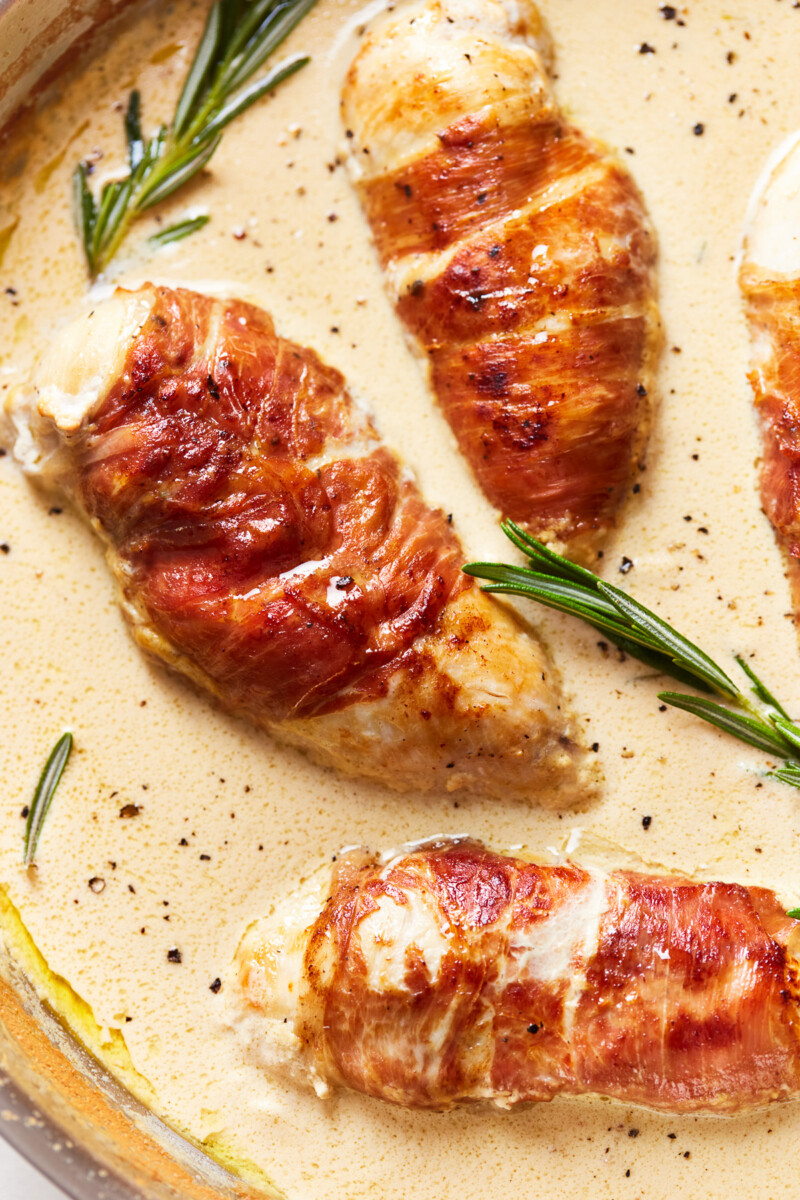 partial overhead view of prosciutto wrapped chicken in a frying pan with rosemary.