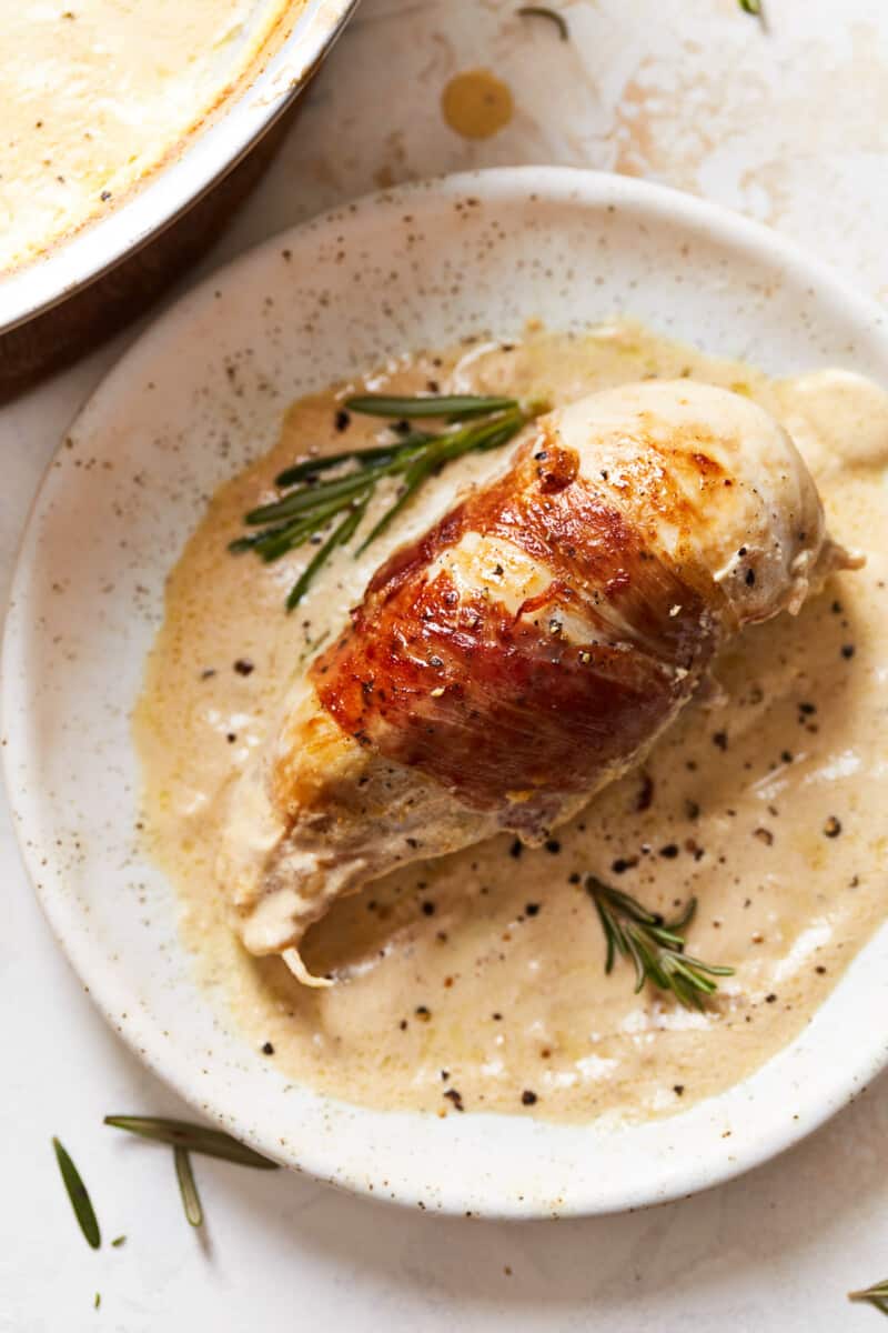 overhead view of a prosciutto wrapped chicken breast on a white plate with sprigs of rosemary.