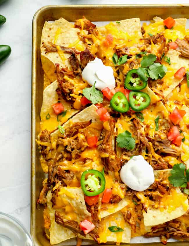 partial overhead view of pulled pork nachos on a baking sheet topped with sour cream, jalapenos, tomatoes, and cilantro.