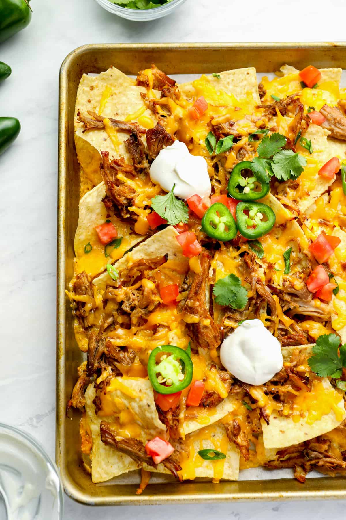 partial overhead view of pulled pork nachos on a baking sheet topped with sour cream, jalapenos, tomatoes, and cilantro.