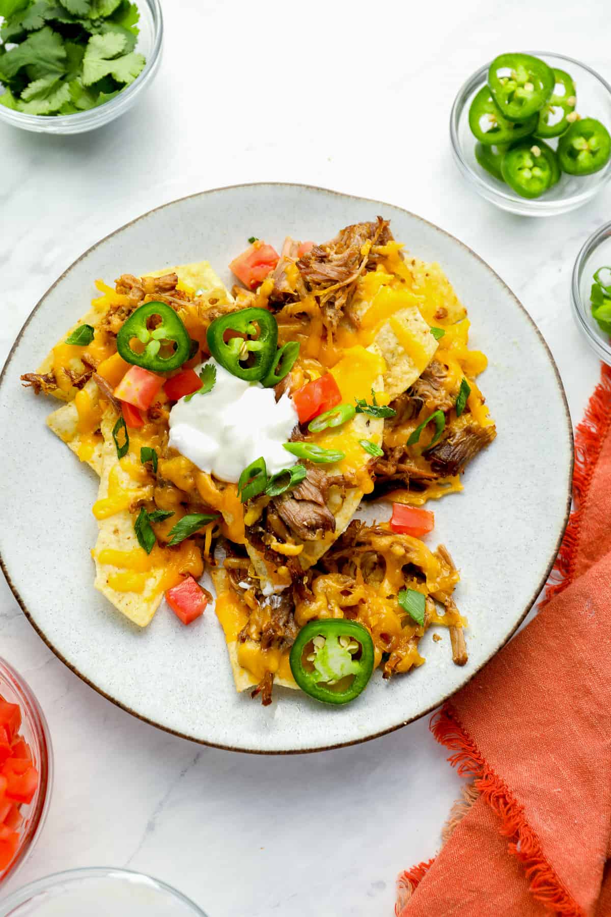 overhead view of a serving of pulled pork nachos on a white plate topped with jalapenos, tomatoes, sour cream, and cilantro.
