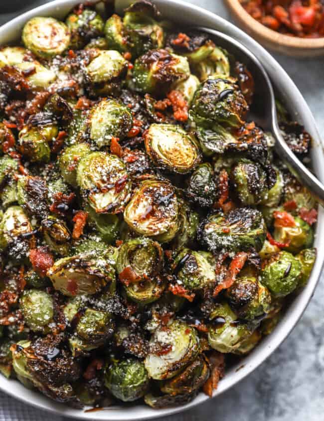 partial overhead view of roasted brussels sprouts topped with bacon, cheese, and balsamic in a serving bowl with a serving spoon.