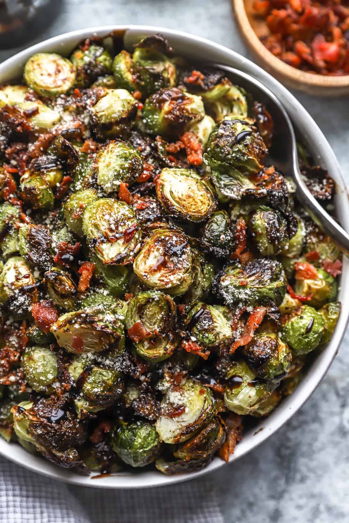partial overhead view of roasted brussels sprouts topped with bacon, cheese, and balsamic in a serving bowl with a serving spoon.