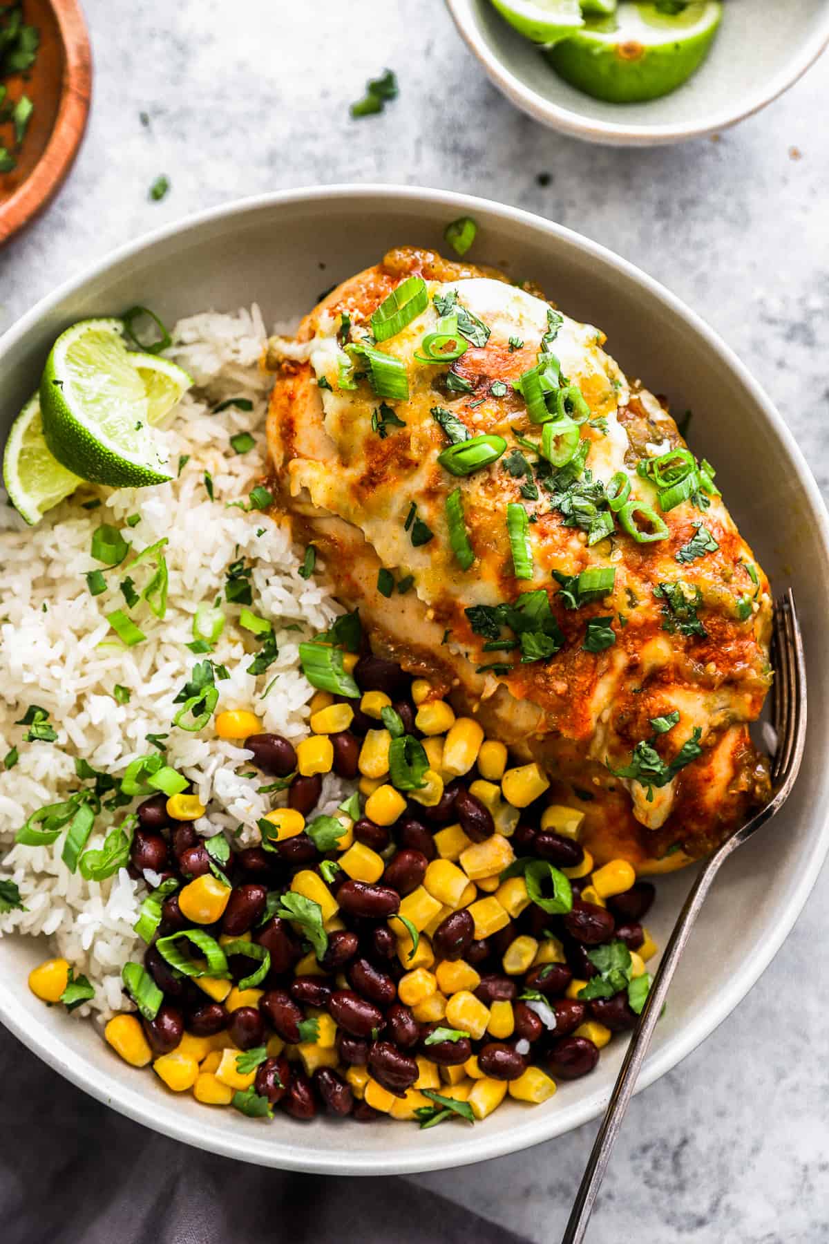 overhead view of a salsa verde chicken breast on a white plate with rice, corn and beans, and lime wedges.
