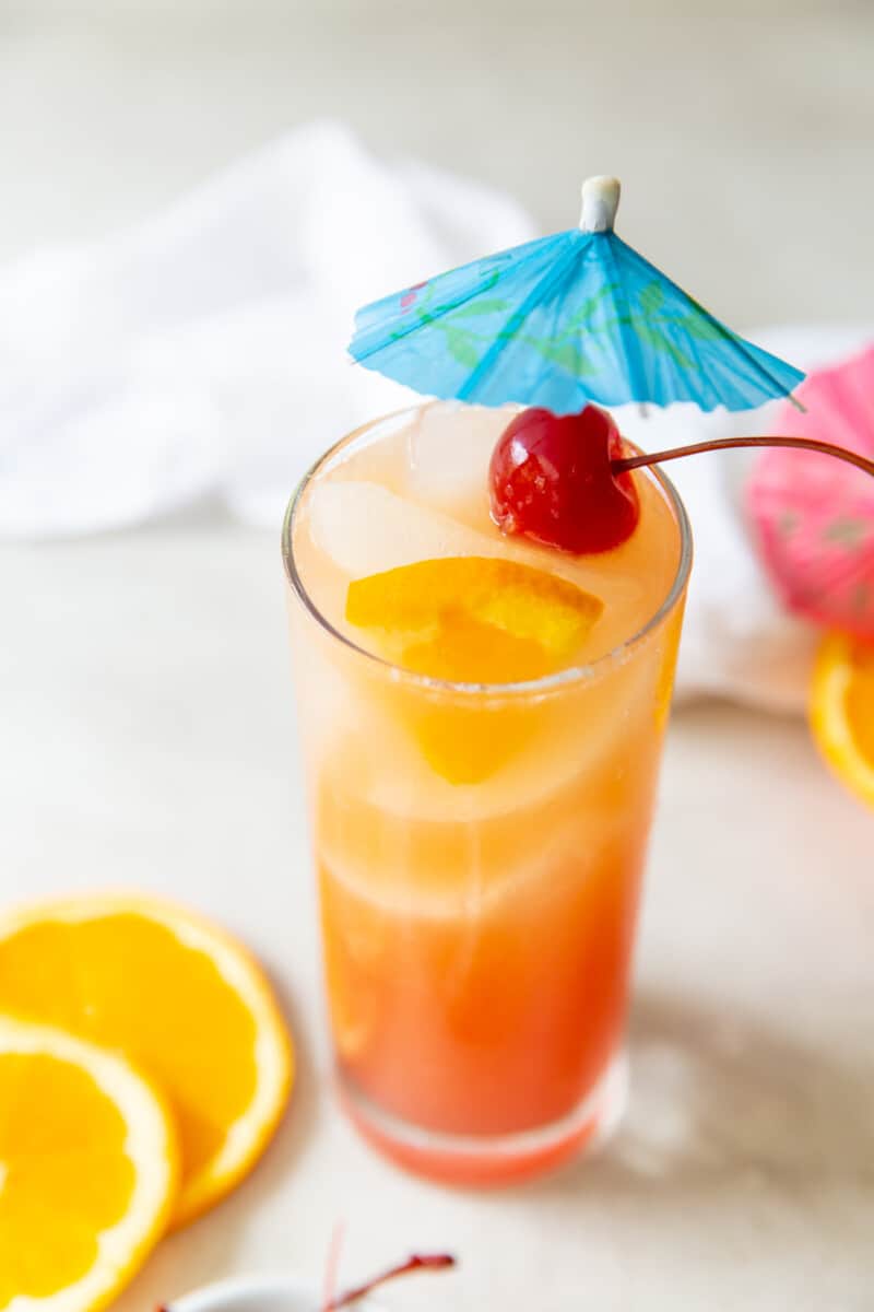 three quarters view of sex on the beach in a tall glass with a cherry skewered on a blue cocktail umbrella.