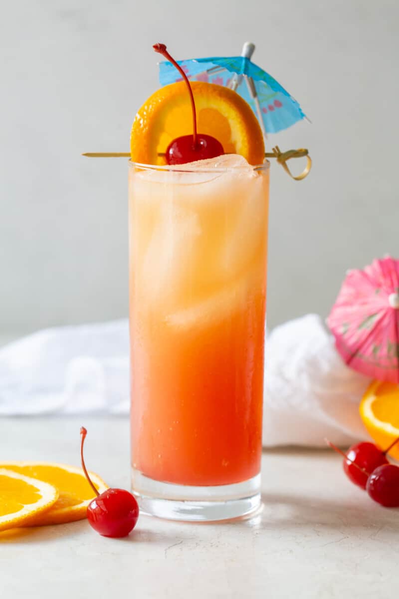 side view of sex on the beach in a tall glass with ice and a cherry, orange slice, and blue cocktail umbrella.