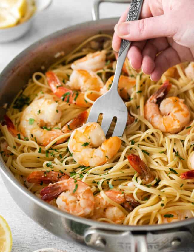 a hand using a slotted spoon to scoop a shrimp from a bed of pasta in a frying pan.
